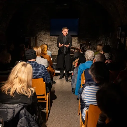 Blick in den voll besetzten Zuschauerraum des Schaffarei Clubs in Feldkirch während einer Vorstellung von „Move fast and break things" von Daniela Egger. Das Publikum sitzt auf Holzstühlen in zwei Reihen zu beiden Seiten eines schmalen Mittelgangs. Am Ende des Gangs steht Schauspieler David Kopp in einem langen schwarzen Mantel und gestikuliert mit beiden Händen. Im Hintergrund ist die beleuchtete Natursteinwand sowie ein dunkler Bildschirm unter dem Gewölbe zu sehen.