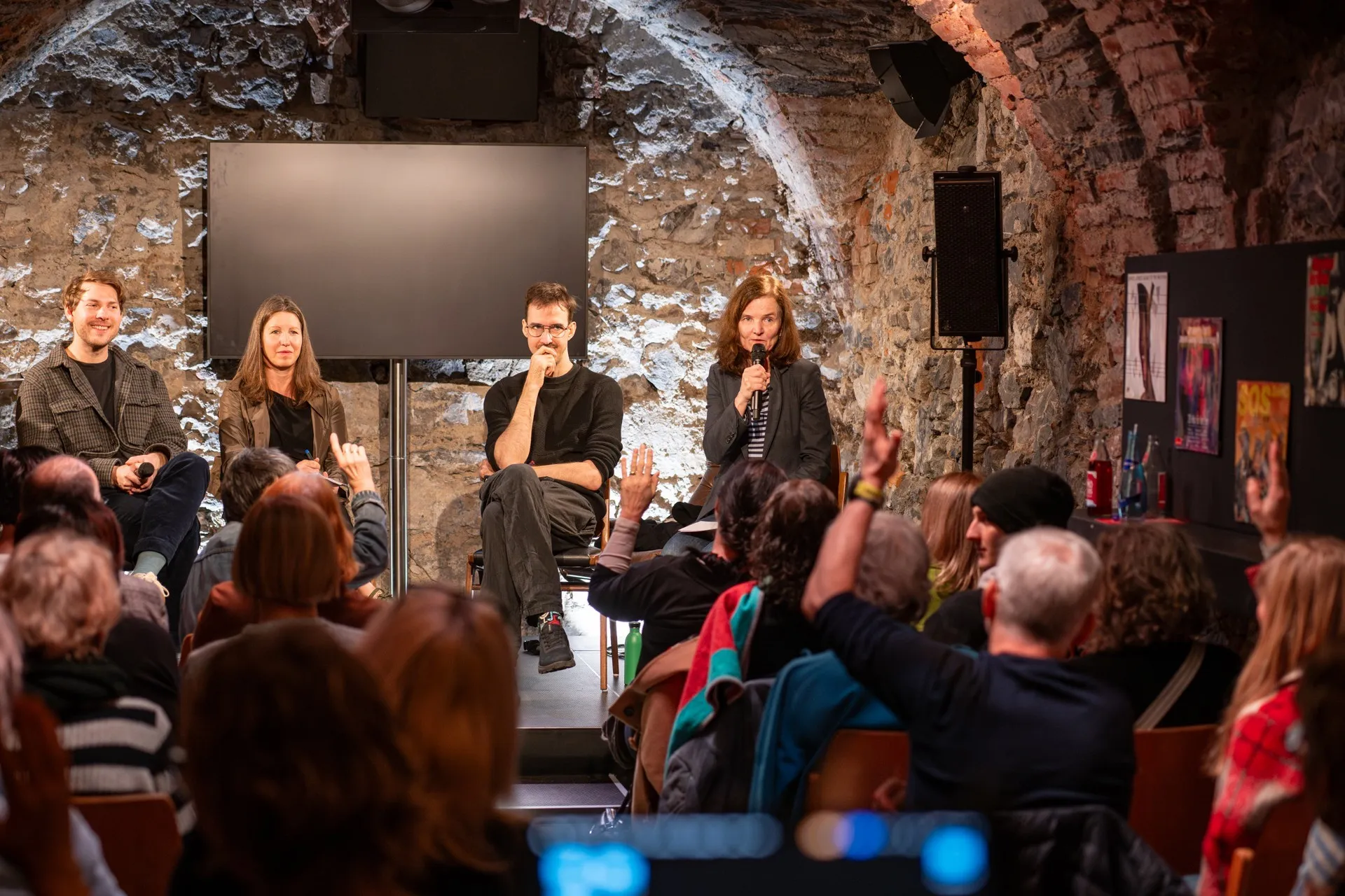 Podiumsdiskussion zum Thema Künstliche Intelligenz im Schaffarei Club in Feldkirch im Anschluss an das Theaterstück „Move fast and break things" von Daniela Egger. Vier Personen sitzen auf Stühlen vor einer Natursteinwand, eine Frau am rechten Rand spricht gerade ins Mikrofon und hält eine Hand erhoben. Links neben ihr sitzt ein Mann mit Brille und nachdenklichem Gesichtsausdruck, daneben eine Frau in olivgrüner Jacke und ganz links ein lächelnder Mann. Im vollbesetzten Zuschauerraum sind mehrere erhobene Hände zu sehen – das Publikum stellt aktiv Fragen. Im Hintergrund hängt ein dunkler Bildschirm an der Natursteinwand.