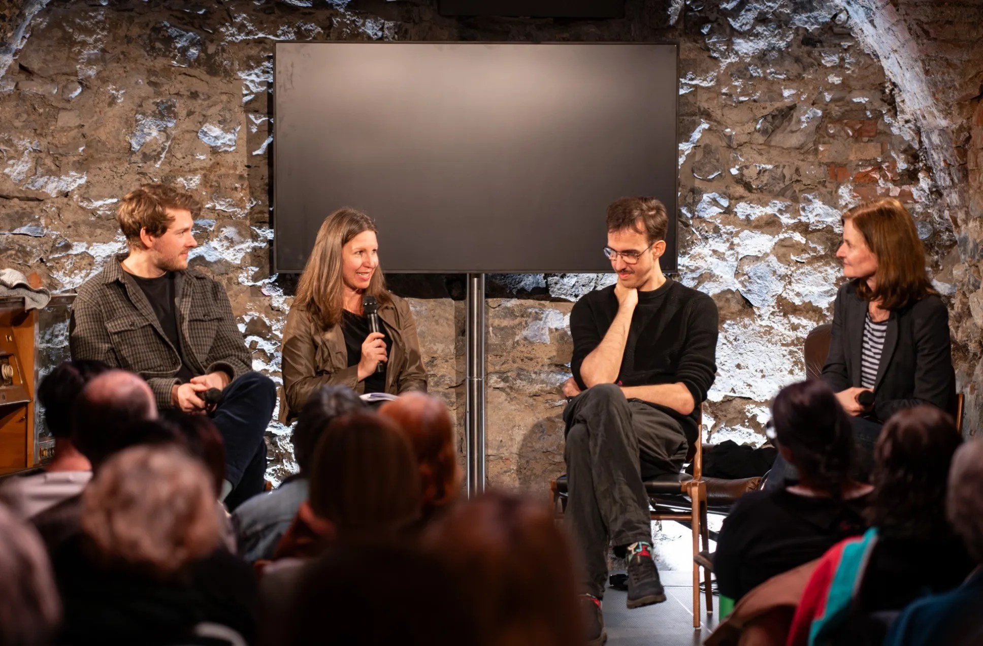 Alt-Text: „Podiumsdiskussion zum Thema Künstliche Intelligenz im Schaffarei Club in Feldkirch im Anschluss an das Theaterstück „Move fast and break things" von Daniela Egger. Vier Personen sitzen auf Stühlen vor einer Natursteinwand und einem leeren Bildschirm. Eine Frau in der Mitte spricht gerade ins Mikrofon. Links neben ihr sitzt ein Mann in kariertem Hemd, rechts von ihr ein Mann mit Brille, der nachdenklich die Hand ans Kinn hält, und ganz rechts eine Frau im Blazer. Im Vordergrund sind die Köpfe des Publikums zu sehen.