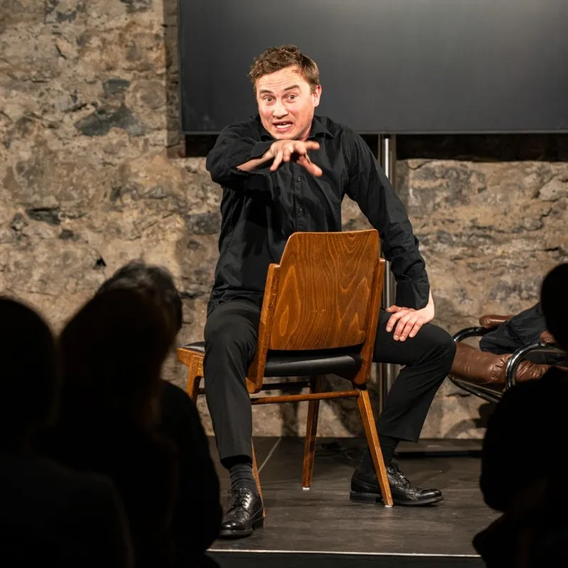 Schauspielszene aus dem Theaterstück „Move fast and break things“ von Daniela Egger, gezeigt im Schaffarei Club in Feldkirch: Ein Schauspieler in schwarzer Kleidung sitzt verkehrt herum auf einem Holzstuhl auf der Bühne und gestikuliert eindringlich mit ausgestreckter Hand ins Publikum. Im Vordergrund sind die Silhouetten der Zuschauer:innen zu sehen, im Hintergrund eine Steinwand, ein Bildschirm und ein Ledersessel.