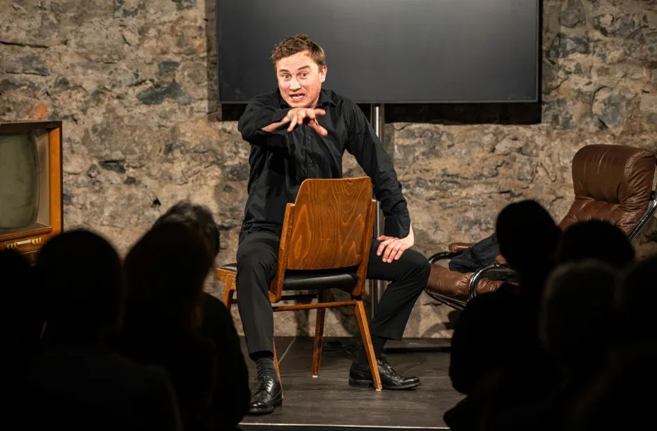 Schauspielszene aus dem Theaterstück „Move fast and break things“ von Daniela Egger, gezeigt im Schaffarei Club in Feldkirch: Ein Schauspieler in schwarzer Kleidung sitzt verkehrt herum auf einem Holzstuhl auf der Bühne und gestikuliert eindringlich mit ausgestreckter Hand ins Publikum. Im Vordergrund sind die Silhouetten der Zuschauer:innen zu sehen, im Hintergrund eine Steinwand, ein Bildschirm und ein Ledersessel.