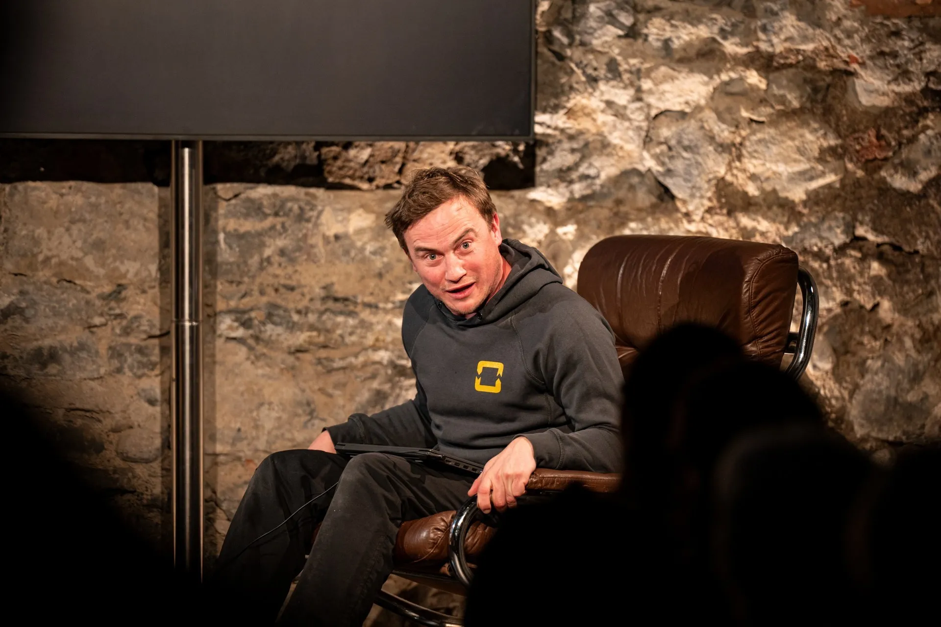 Alt-Text: „Schauspieler David Kopp sitzt in einem braunen Ledersessel auf der Bühne des Schaffarei Clubs in Feldkirch während einer Vorstellung von „Move fast and break things" von Daniela Egger. Er trägt einen dunkelgrauen Hoodie mit gelbem Logo und hält einen Laptop auf dem Schoß. Sein Gesicht ist dem Publikum zugewandt, der Mund leicht geöffnet und die Augen weit aufgerissen – als ob er gerade etwas Überraschendes sagt oder erlebt. Im Hintergrund ist die beleuchtete Natursteinwand sowie ein dunkler Bildschirm zu sehen, im Vordergrund die Silhouetten von Zuschauern.