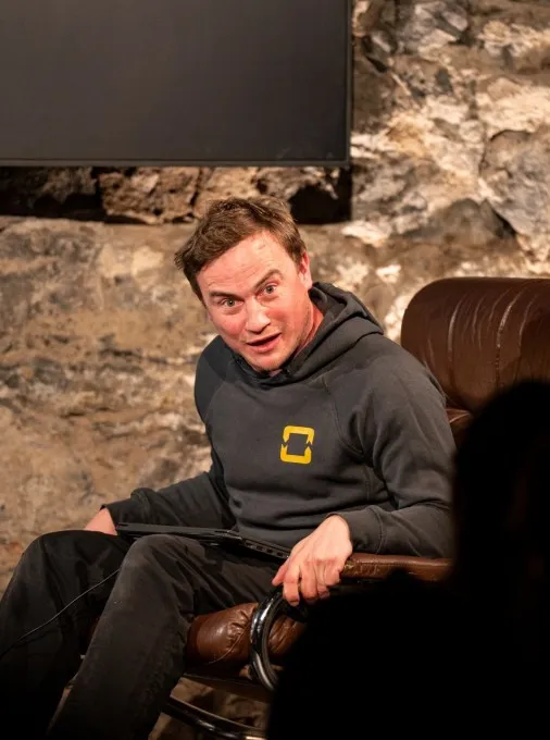 Alt-Text: „Schauspieler David Kopp sitzt in einem braunen Ledersessel auf der Bühne des Schaffarei Clubs in Feldkirch während einer Vorstellung von „Move fast and break things" von Daniela Egger. Er trägt einen dunkelgrauen Hoodie mit gelbem Logo und hält einen Laptop auf dem Schoß. Sein Gesicht ist dem Publikum zugewandt, der Mund leicht geöffnet und die Augen weit aufgerissen – als ob er gerade etwas Überraschendes sagt oder erlebt. Im Hintergrund ist die beleuchtete Natursteinwand sowie ein dunkler Bildschirm zu sehen, im Vordergrund die Silhouetten von Zuschauern.