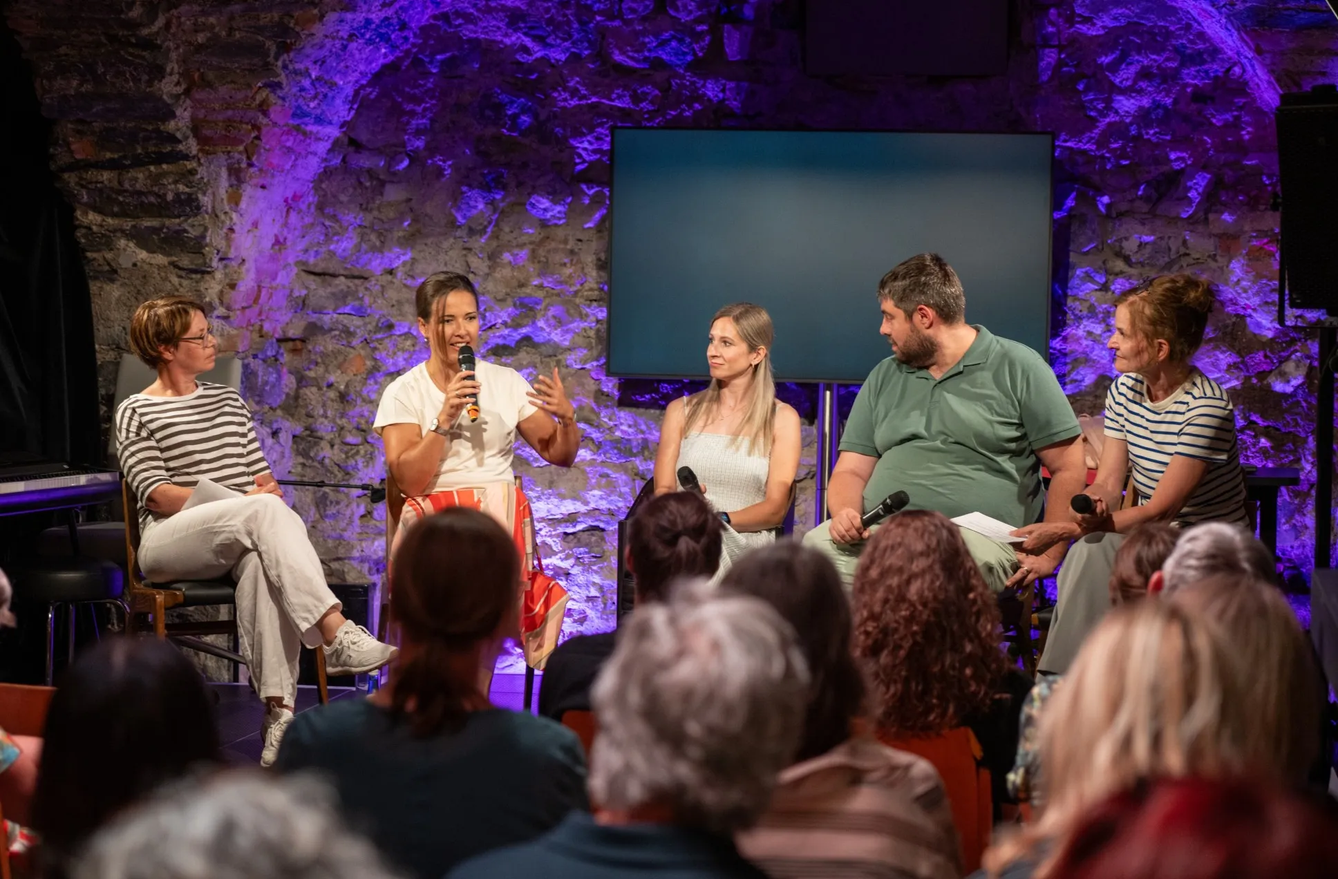 Eine Podiumsdiskussion mit fünf Teilnehmenden in einem rustikalen Gewölbekeller. Die Teilnehmer sitzen auf Stühlen auf einer kleinen Bühne, die von lilafarbenem Licht beleuchtet wird. Im Hintergrund ist eine alte Steinwand und ein großer, dunkler Bildschirm zu sehen. Eine Frau mit kurzem Haar und einem weiß-orange-gestreiften Rock spricht in ein Mikrofon und gestikuliert. Links neben ihr sitzt eine Frau im Querstreifen-Shirt, rechts sitzen zwei weitere Frauen und ein Mann, die ihr zuhören. Der Mann trägt ein grünes Poloshirt und hält ein Mikrofon. Im Vordergrund sind die Hinterköpfe und Schultern des Publikums zu sehen.
