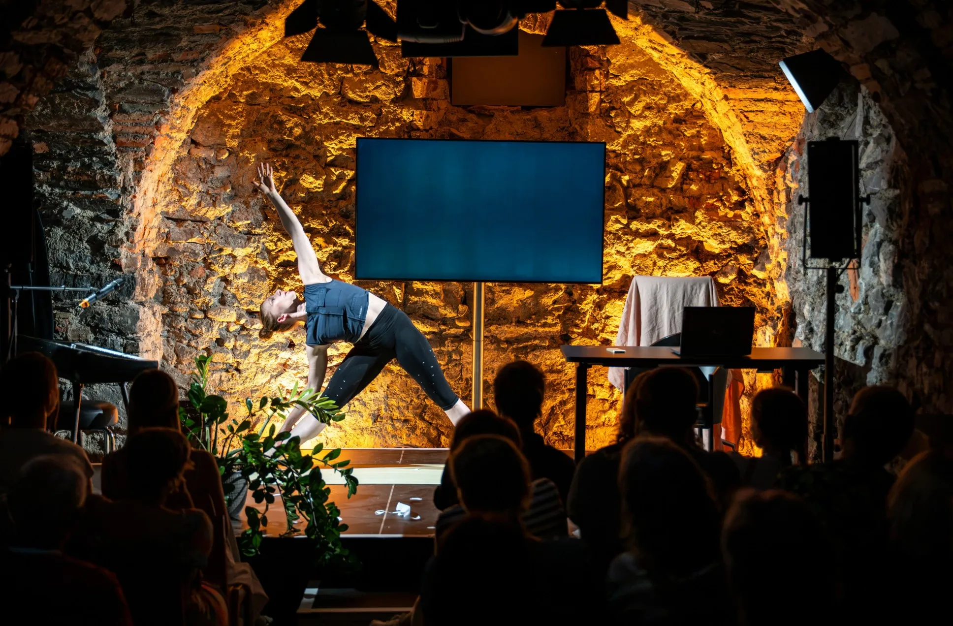 Schauspielerin Vivien Causemann macht eine Yoga-Geste auf einer Bühne vor einer beleuchteten Natursteinwand. Im Hintergrund stehen ein Flachbildschirm und ein Schreibtisch, im Vordergrund sitzt das Publikum als Silhouette.