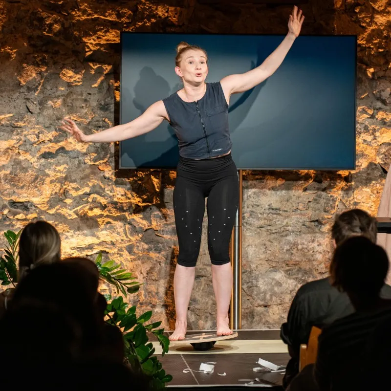 Schauspielerin Vivien Causemann steht barfuß auf einem Balance-Board auf einer Bühne und spricht mit erhobenen, ausgestreckten Armen zum Publikum. Sie trägt dunkle Sportkleidung, im Hintergrund ist eine rustikale Steinwand mit Flachbildschirm zu sehen.