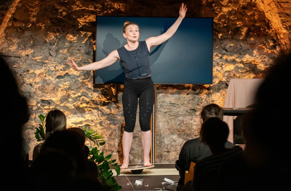 Schauspielerin Vivien Causemann steht barfuß auf einem Balance-Board auf einer Bühne und spricht mit erhobenen, ausgestreckten Armen zum Publikum. Sie trägt dunkle Sportkleidung, im Hintergrund ist eine rustikale Steinwand mit Flachbildschirm zu sehen.
