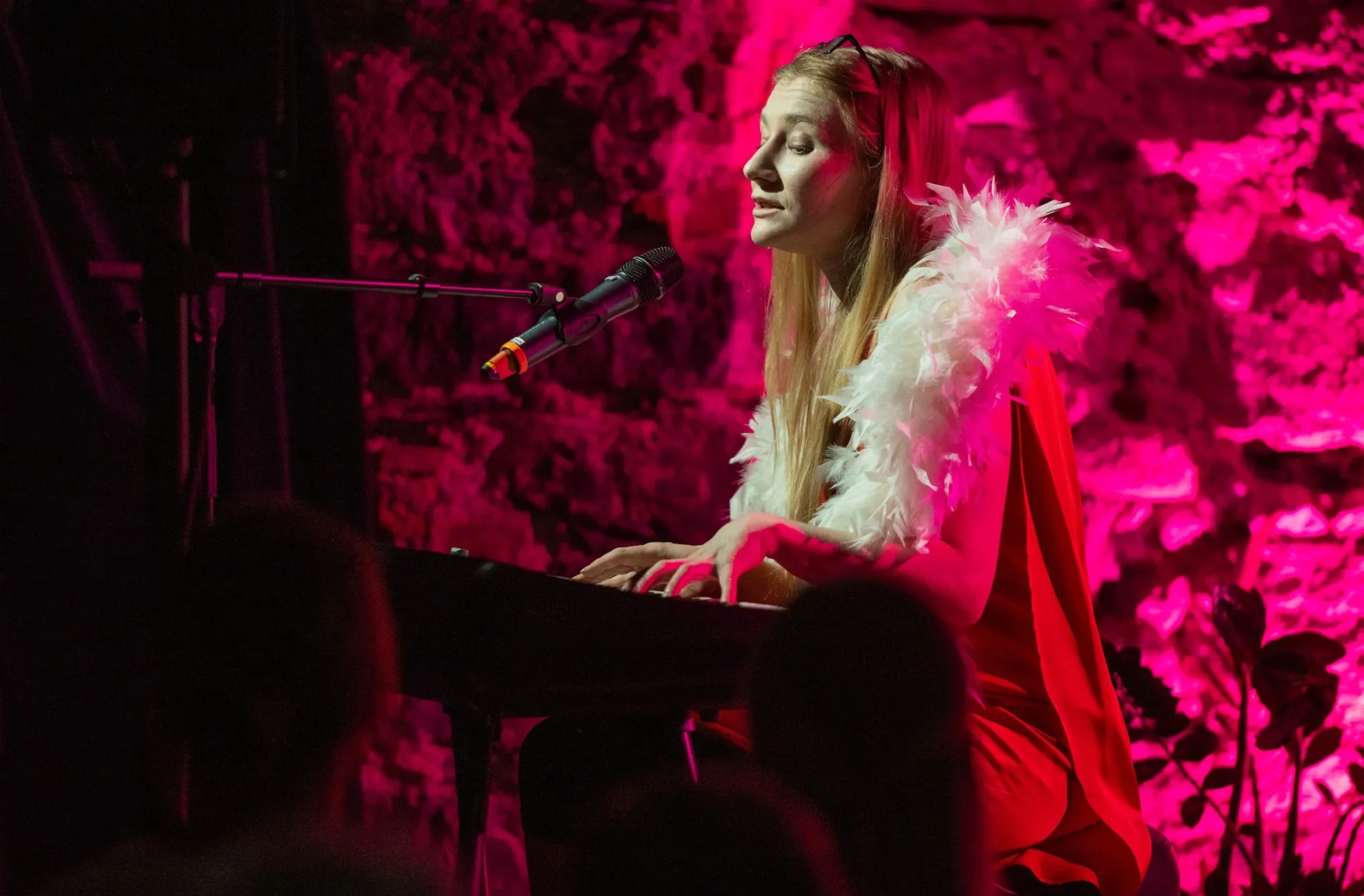 Schauspielerin Vivien Causemann sitzt im Profil an einem Keyboard und singt in ein Mikrofon. Sie trägt ein rotes Kleid und eine weiße Federboa. Der Hintergrund ist in auffälliges pinkes Licht getaucht.